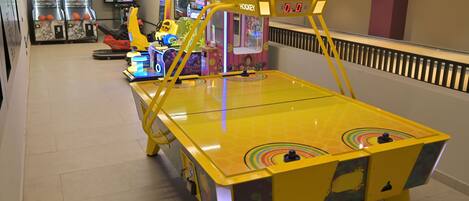 Um salão de jogos coberto, bem iluminado, com diversos jogos eletrônicos, incluindo uma mesa de pingue-pongue amarela e uma máquina de hóquei.