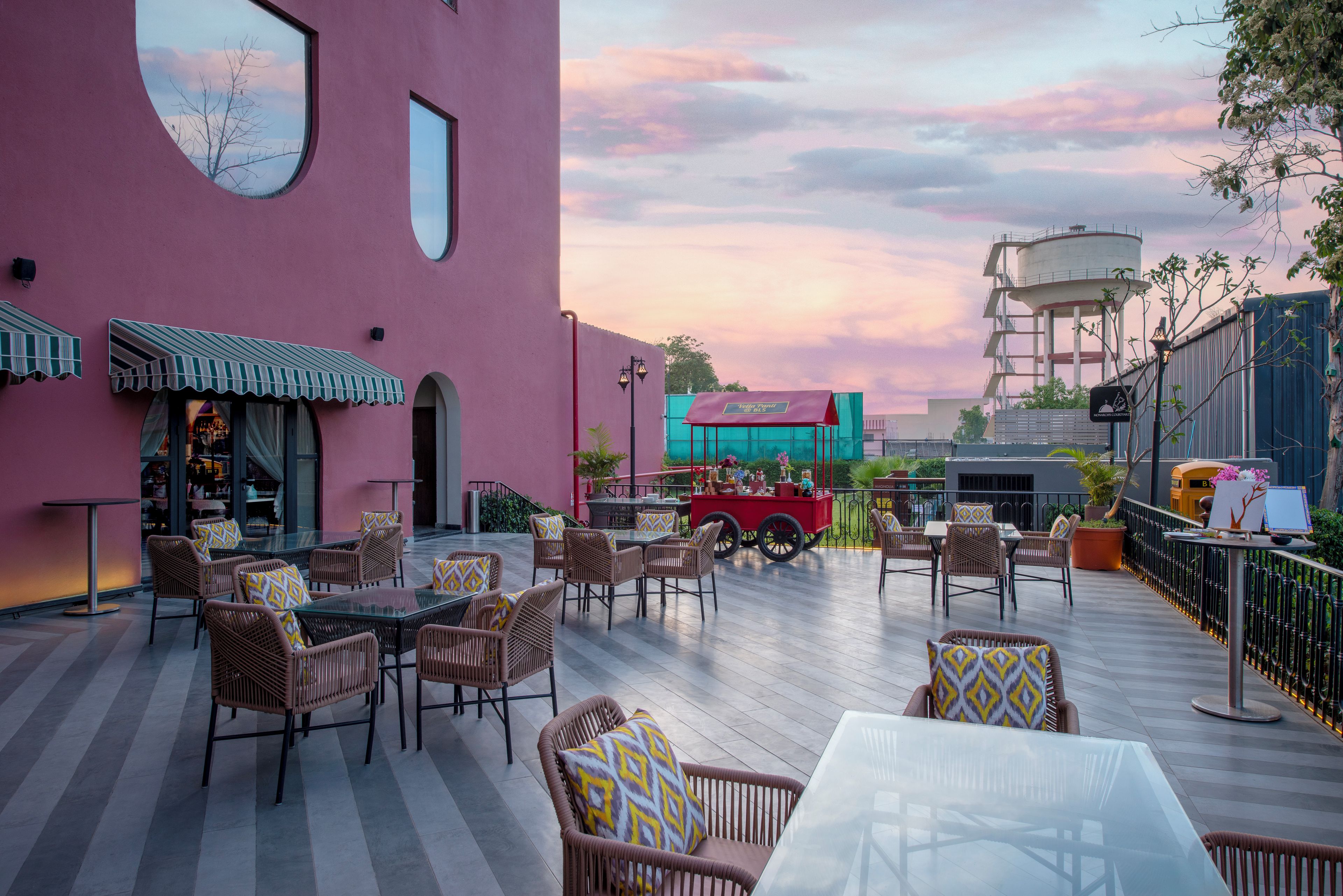 An outdoor seating area with wicker furniture and a glass table, a pink building with large windows, and a sunset sky.