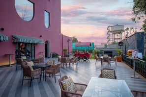 Un área de estar al aire libre con muebles de mimbre y una mesa de vidrio, un edificio rosa con ventanas amplias y un cielo al atardecer.