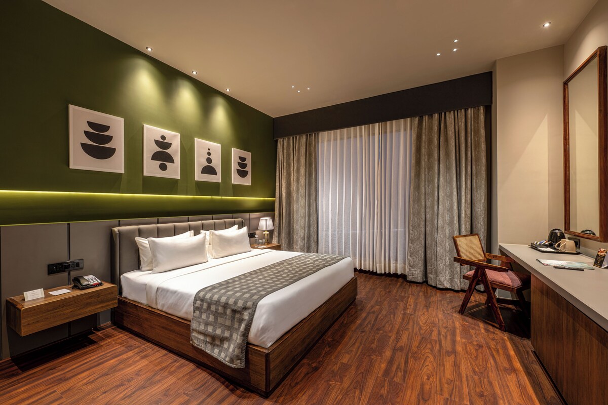 A modern hotel room with a large bed, a desk with a chair, a bedside table, and a large window with curtains.