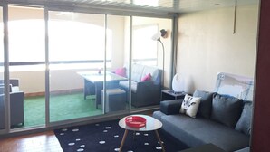 Un salon moderne avec une porte-fenêtre coulissante donnant sur un balcon, un canapé gris, une petite table ronde sur laquelle se trouve un objet rouge, et un lampadaire.