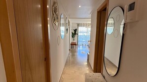 Un pasillo con puertas de madera, suelo de mármol y un banco. Hay espejos en la pared derecha y un teléfono.