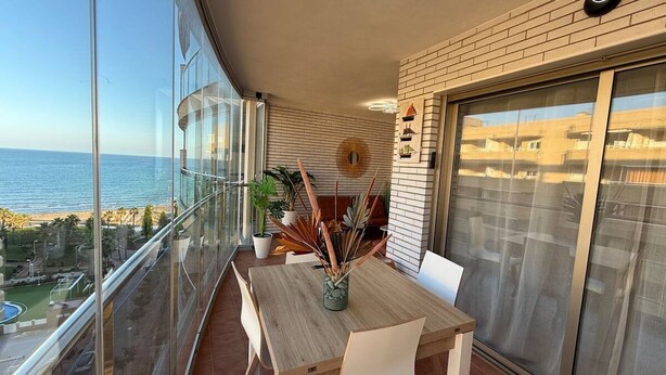 Un balcón con mesa y sillas de madera, plantas en macetas y vista al mar.