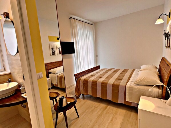 A hotel room with a bed, bedside table, lamp, and a bathroom with a sink and mirror. - Residenza Delfina (Canale Monterano)