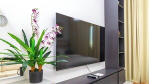 A modern living room with a large flat-screen TV mounted on a dark wall, a potted plant with green leaves and purple flowers, and a beige sofa. - Bloomfields Lavish STU in Oasis Masdar (Abu Dhabi)
