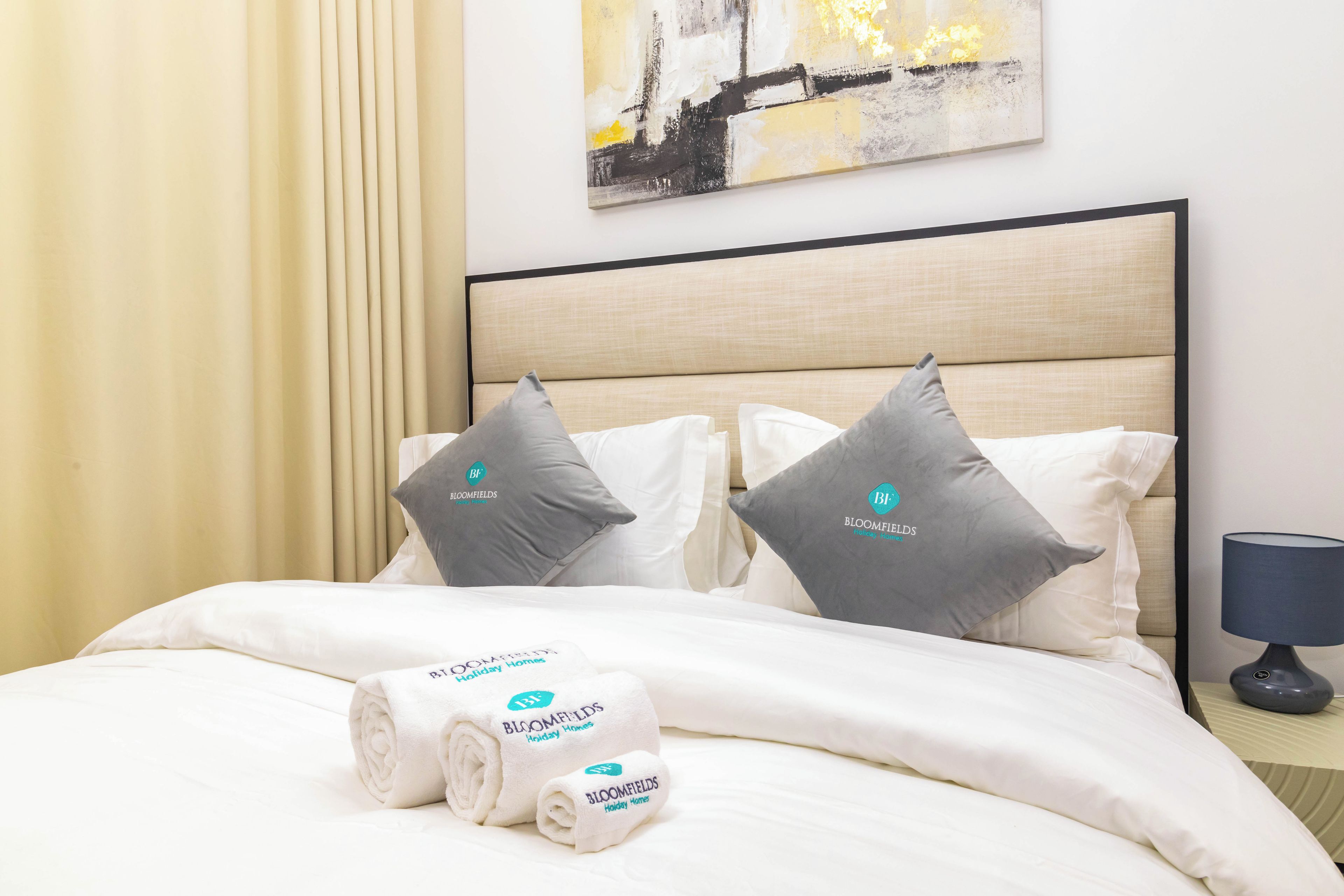 A neatly made bed with white linens and two gray pillows, a rolled white towel, and a bedside table with a lamp.