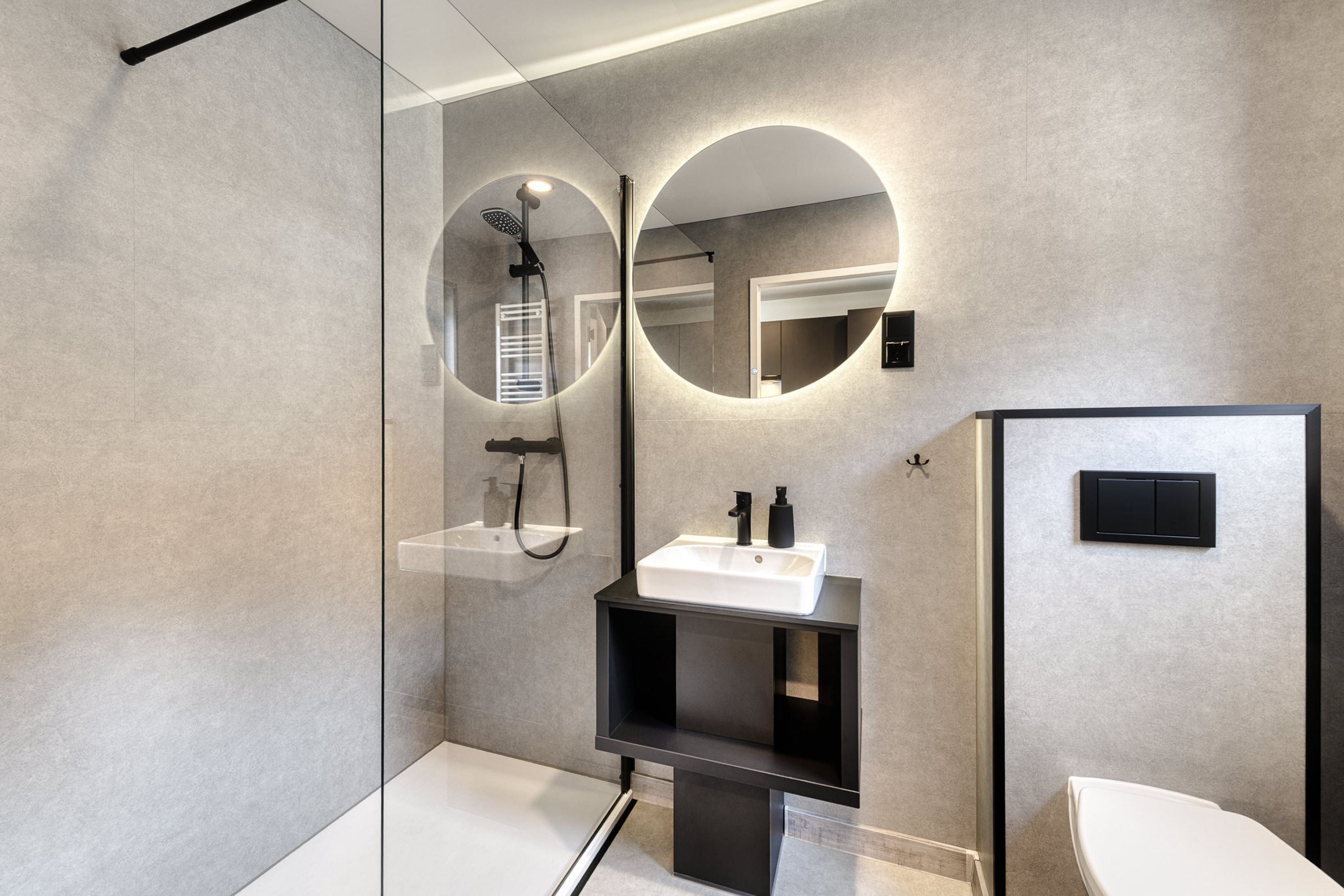 A modern bathroom with a glass shower enclosure, a white sink with black fixtures, and a round mirror with a circular light.