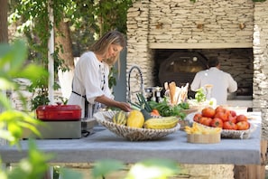 An outdoor cooking area with a stone oven, a table with fresh produce, and a person preparing food. - Votsalo Sea Side (Andros)