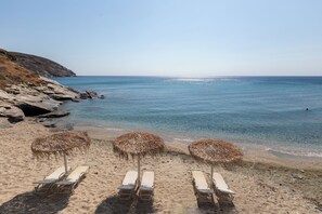 A beach with clear water, rocky shoreline, and sun loungers with thatched umbrellas. - Votsalo Sea Side (Andros)