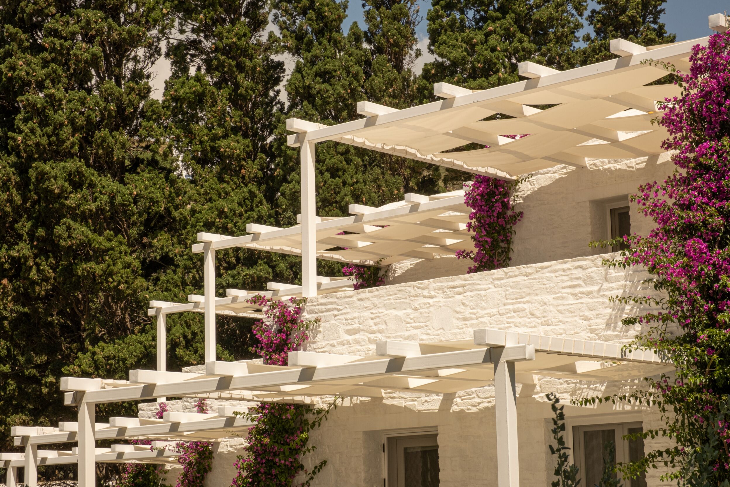 A white pergola structure with a stone building in the background, surrounded by lush greenery and flowering plants.