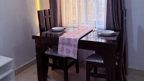 A dining area with a table set for two, a window with curtains, and a wall-mounted light. - NYALI 1BR/Hot Shower/AC/Parking behind Quickmart (Mombasa)