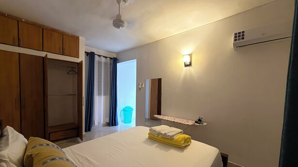 A hotel room with a bed, a wardrobe, a mirror, an ironing board, and an air conditioning unit. - NYALI 1BR/Hot Shower/AC/Parking behind Quickmart (Mombasa)