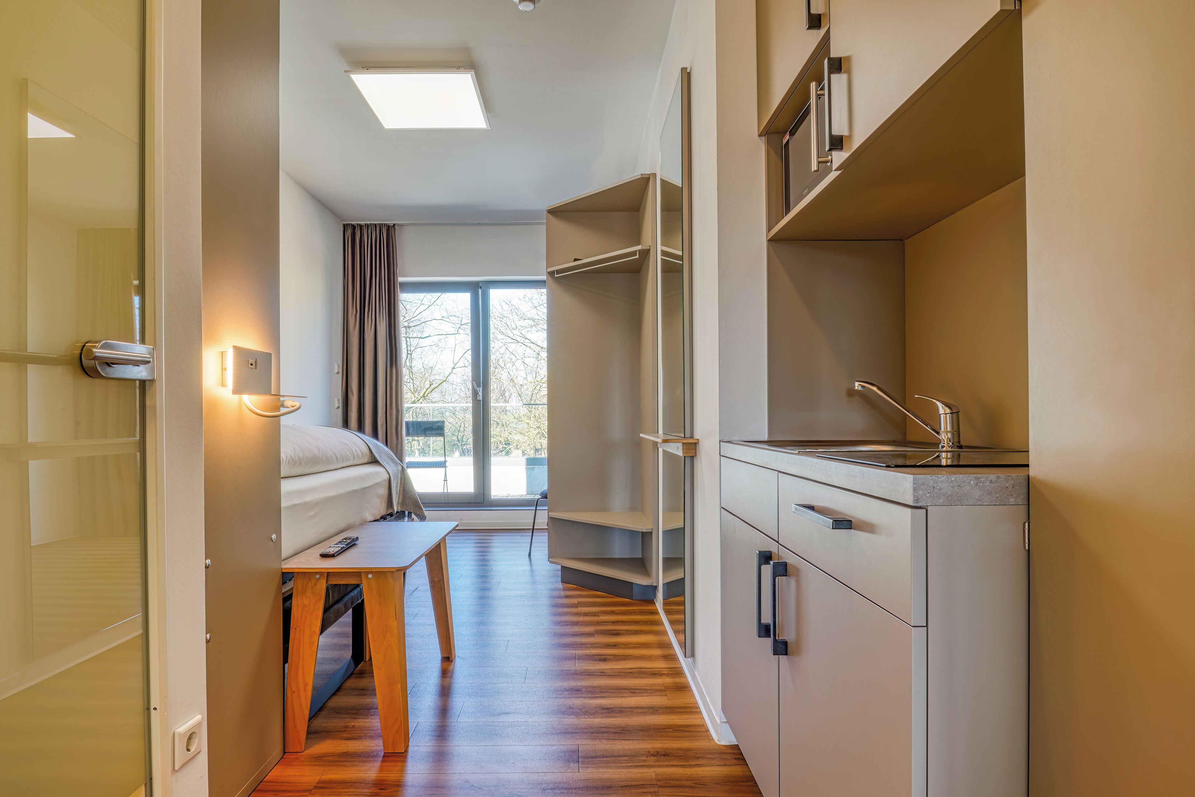 Habitación de hotel pequeña y funcional, con cama, una mesita, kitchenette y puerta corredera que da a un balcón.