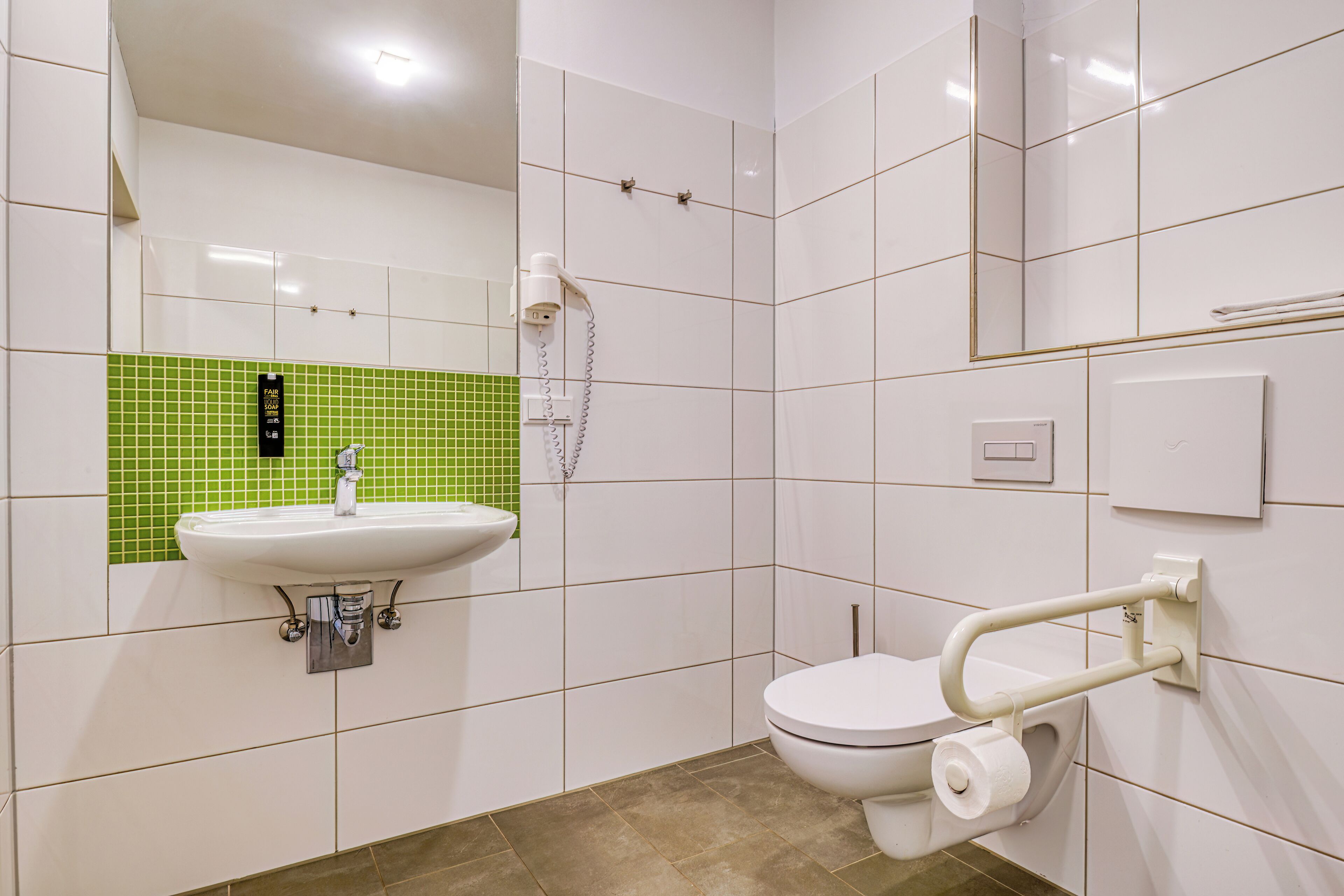 A bathroom with a white sink, green-tiled wall, white toilet with a handrail, and a mirror.