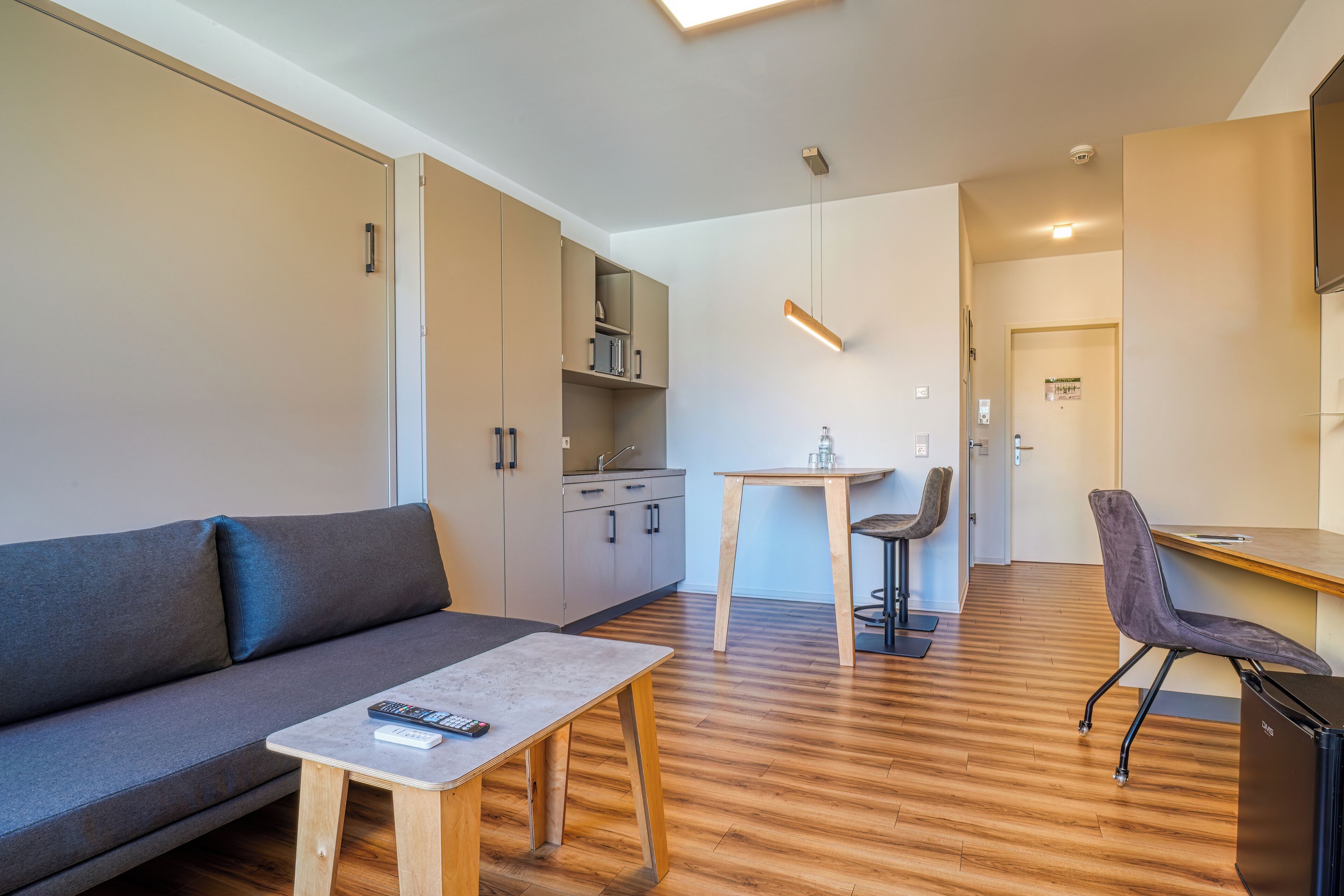 A modern living room with a sofa, a coffee table, a dining table with chairs, and a kitchen area with cabinets and a hanging light fixture.