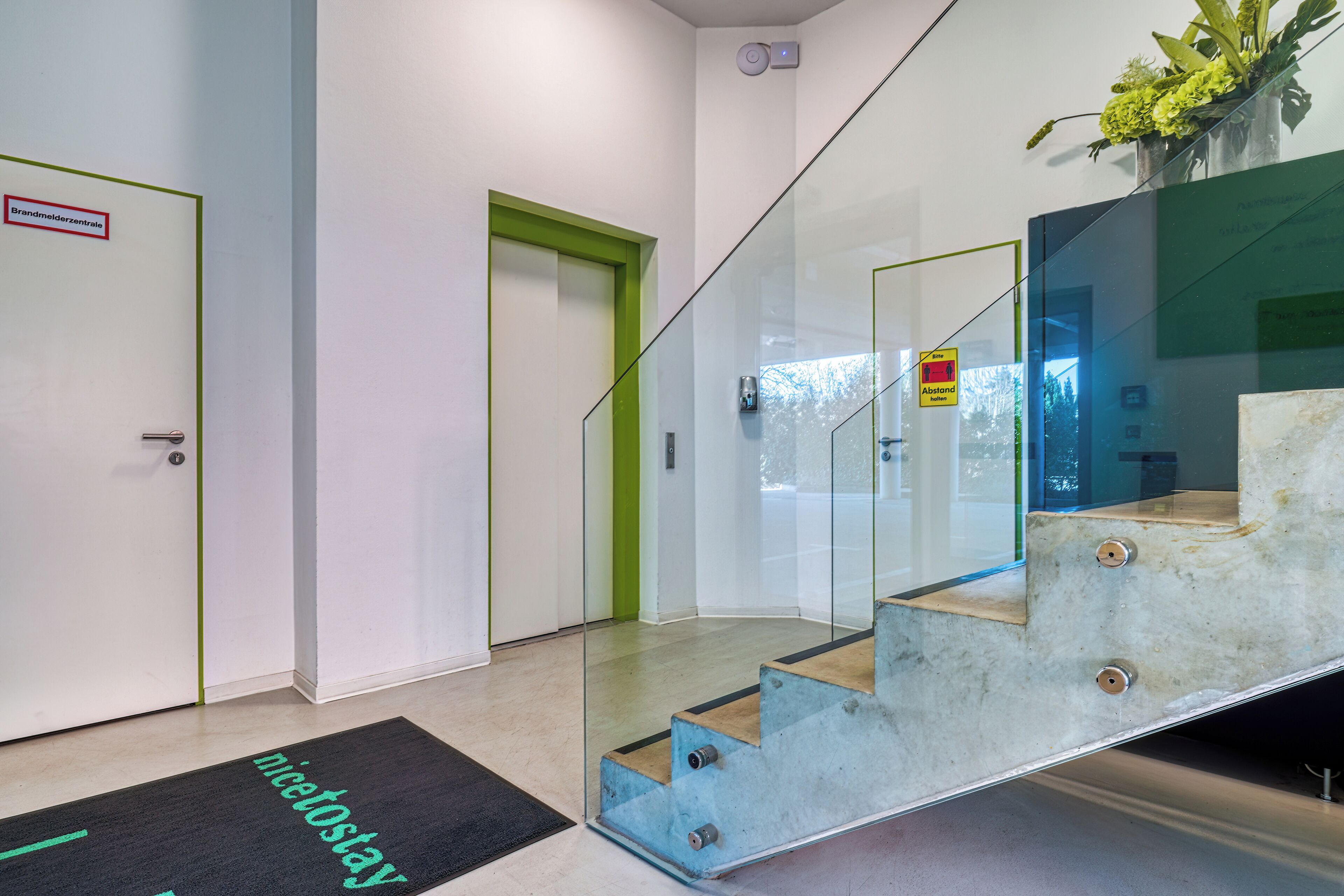 Ein modernes Interieur mit einer Glastreppe, einer Fußmatte mit dem Aufdruck "nicetostay" und einer Tür mit der Beschriftung "Brandmelderszentrum".