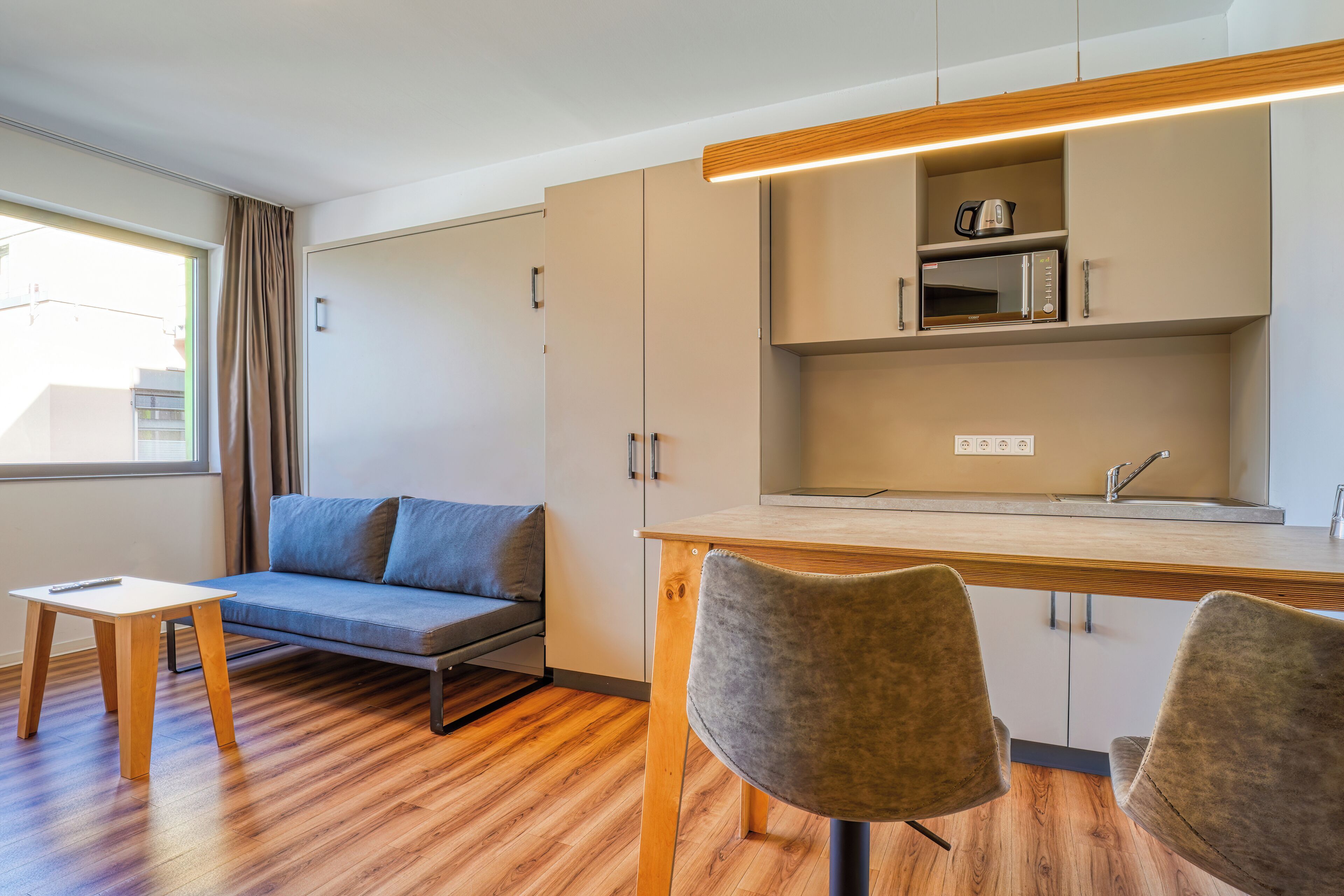 A modern kitchenette with a built-in microwave and kettle, a wooden dining table, and a sofa.