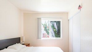 Een hotelkamer met een bed, nachtkastje, lamp, raam met uitzicht op bomen en een gebouw, en een Amerikaanse vlag aan de muur.