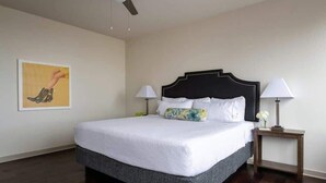 Una cama bien hecha con sábanas blancas y un cabecero oscuro, dos lámparas de noche, una mesita con una lámpara y un cuadro en la pared.