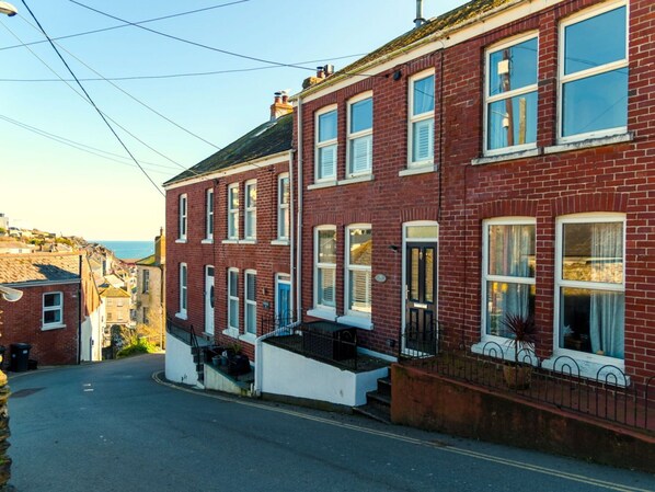A row of red brick townhouses with white window frames and shutters, a narrow street, and a clear view of the sea in the background.