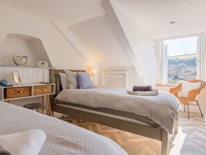 A bedroom with a loft-style ceiling, a bed with a grey duvet, a wicker side table, a chair, and a view of the countryside through a window.