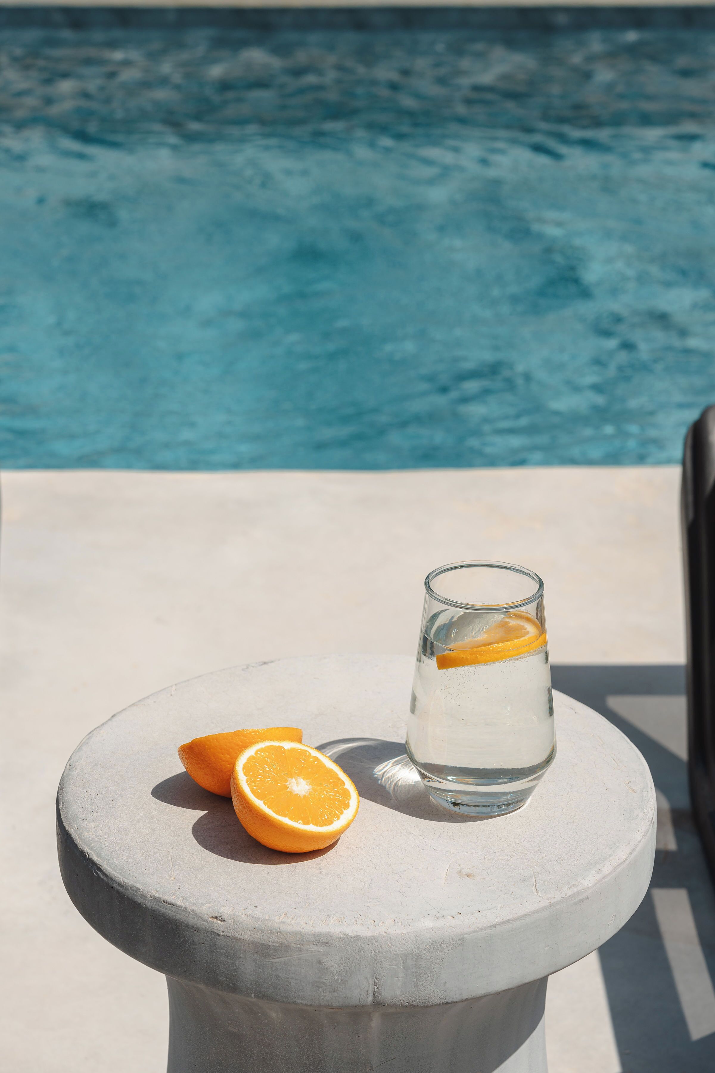 Un verre d’eau avec une tranche d’orange, posé sur une table en béton, près d’une piscine.
