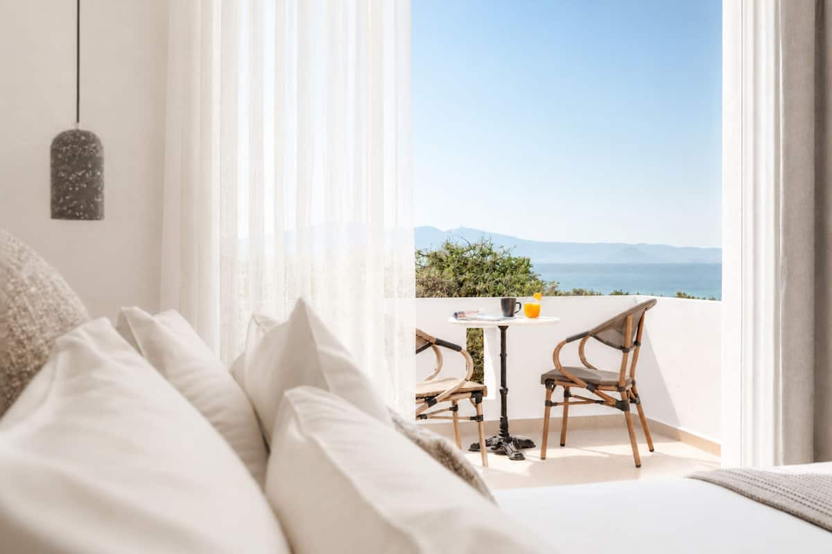 A bedroom with a large window, a bed with white bedding, and a view of the sea.