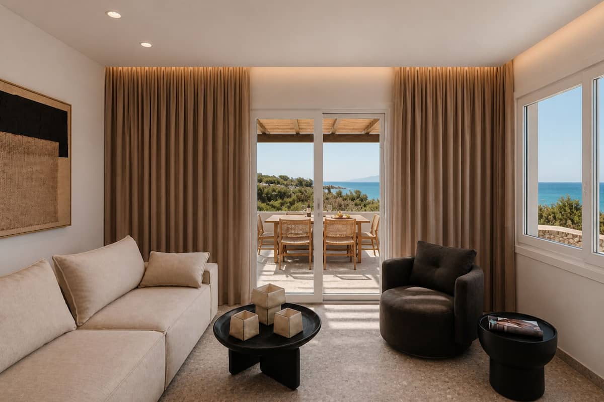 A modern living room with a sofa, armchair, and coffee table, a balcony with outdoor furniture, and a view of the sea.