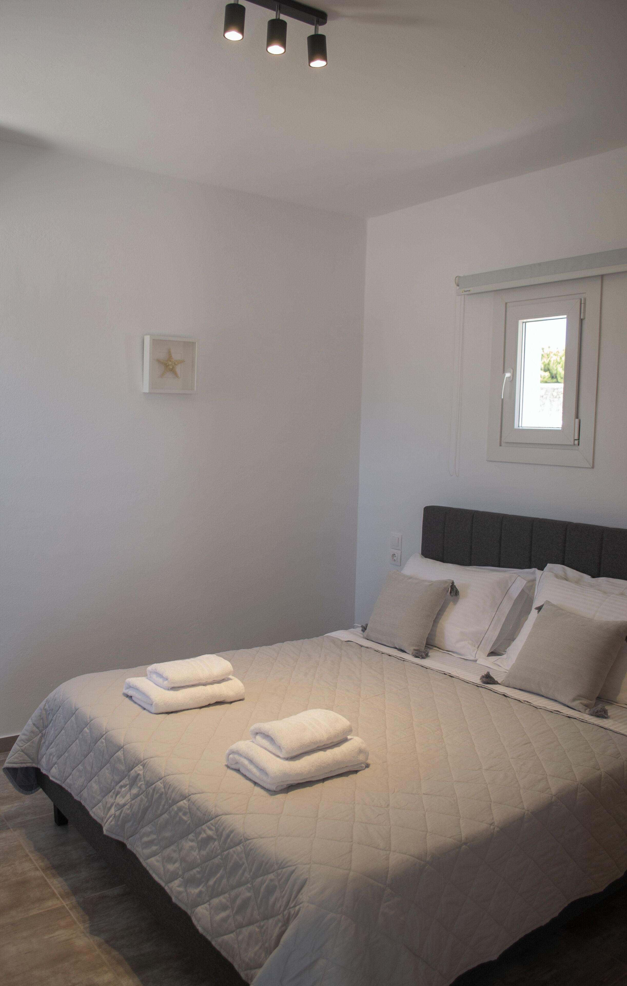Una cama bien hecha con cubrecama acolchada y dos toallas dobladas, un cuadro en la pared, y una ventana con vistas a zonas verdes.