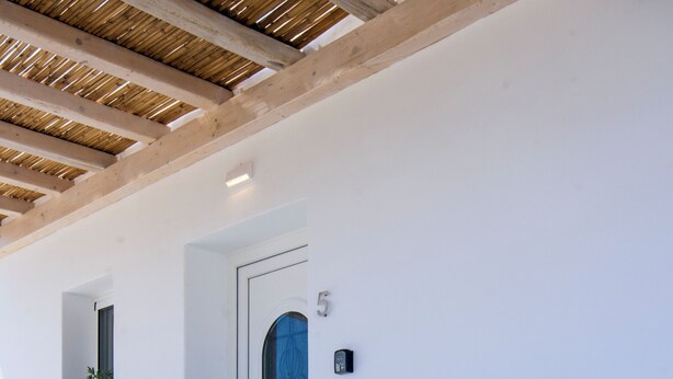 Una habitación con paredes blancas, un banco de madera y dos sillas blancas acolchadas, una pequeña mesa con una planta, y una puerta marcada con el número 5.