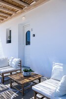 Una habitación con paredes blancas, un banco de madera y dos sillas blancas acolchadas, una pequeña mesa con una planta, y una puerta marcada con el número 5.
