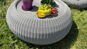 Una mesa redonda de mimbre con una planta en maceta, un vaso de bebida verde, un cuenco de fruta y un par de zapatillas.