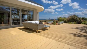A modern outdoor deck with a sofa and chairs, a dining area with tables, and a view of the ocean.