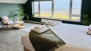 A bedroom with a large window, a bed with a green throw, a chair, a small table, and a plant.