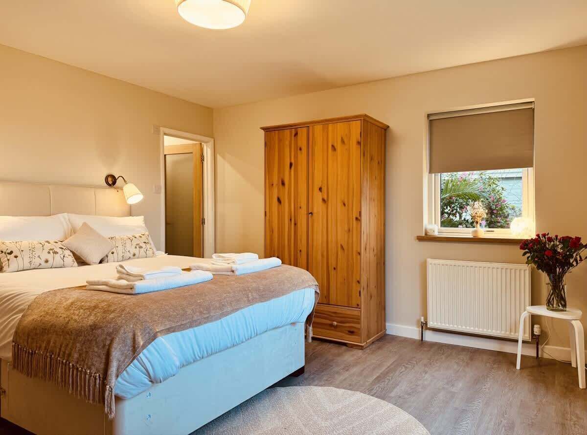 Un dormitorio con cama, armario de madera, radiador, ventana con vistas y mesita de noche con un florero.