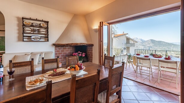 A dining area with a brick fireplace, a table set for four, and a view of mountains through large windows. - Villa Ruju in Dorgali (Dorgali)
