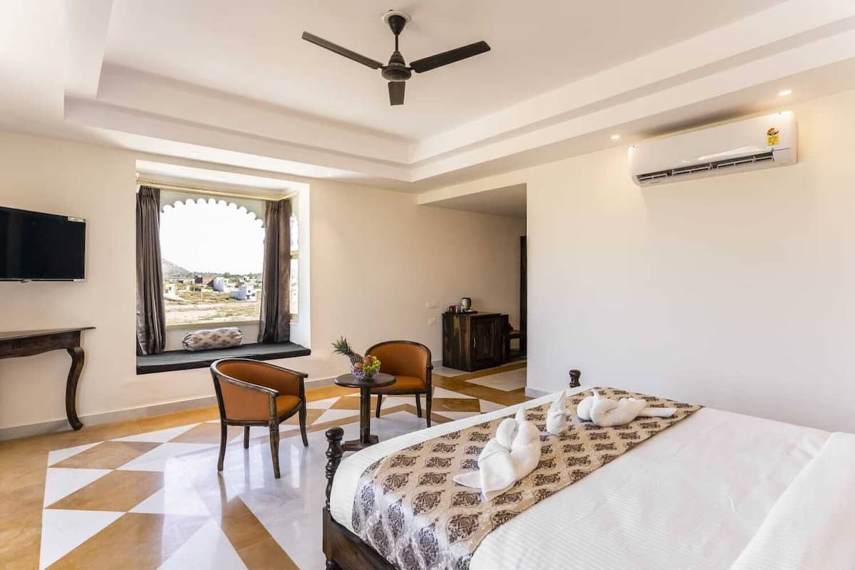 Habitación de hotel con cama, dos sillas, una mesita, televisor, ventana con cortinas y equipo de aire acondicionado.