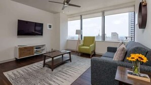 A modern living room with a flat-screen TV, a ceiling fan, a sofa, a coffee table, a green armchair, and a large window with a city view. - Stunning CozySuites on Elm St. 11 (Dallas)