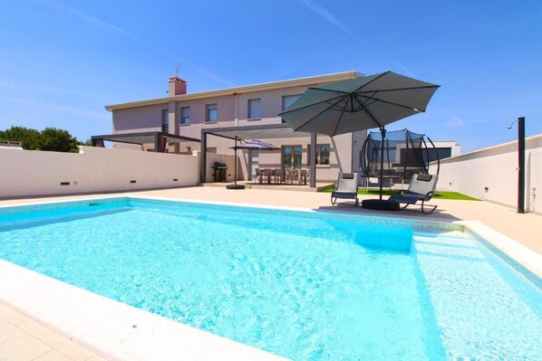 Una piscina rettangolare con acqua cristallina e blu, circondata da un solarium piastrellato. Si vedono lettini da relax e un ombrellone che offre ombra.