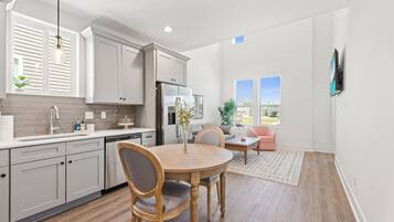 Une cuisine moderne avec des meubles blancs, une table à manger avec des chaises, un réfrigérateur et une fenêtre avec des stores.