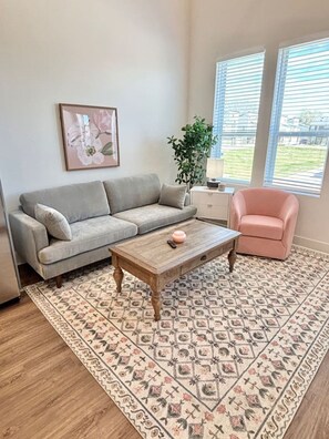 A living room with a grey sofa, a wooden coffee table, a pink chair, a patterned rug, and a plant.
