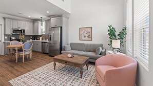 Une cuisine moderne équipée d’appareils électroménagers en acier inoxydable, un coin repas avec une table et des chaises en bois, un canapé gris et un fauteuil rose.