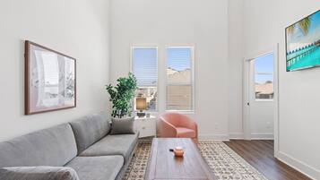 Un salon spacieux avec un canapé gris, une table basse en bois et un fauteuil rose. Il y a une grande fenêtre avec des stores, une plante en pot et un tableau encadré au mur.
