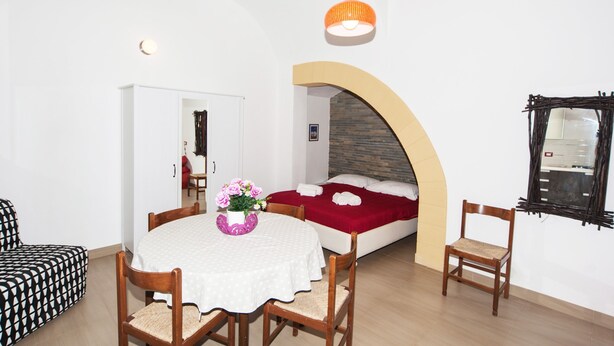 Una habitación con una cama, una mesa de comedor con sillas, una mesita con un jarrón de flores y una lámpara colgante.