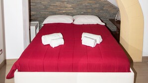 Una cama con colcha roja y almohadas blancas, cabecera de ladrillo y mesita de noche blanca con una lámpara.