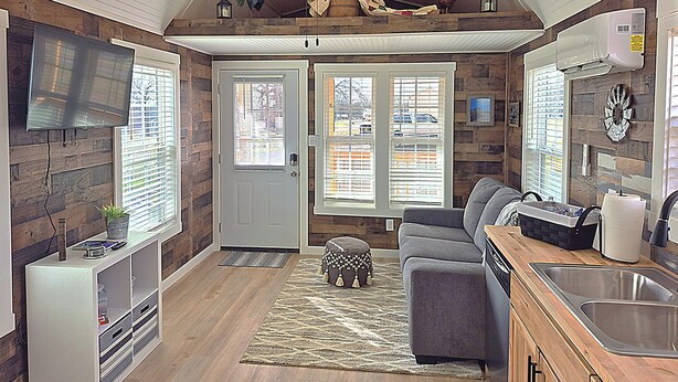 A compact kitchen with a built-in oven, a sink, and a dining area with a grey sofa and a small table. - Redbud Tiny Home Resort (Big Cabin)