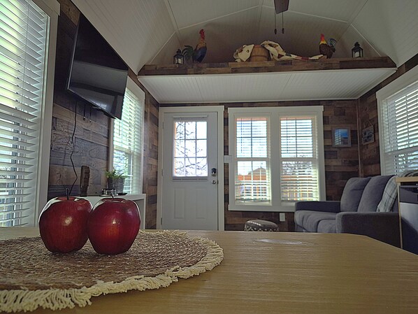 A dining area with a wooden table, two apples, and a woven placemat. There are two windows with blinds, a door, and a stone wall. - Redbud Tiny Home Resort (Big Cabin)