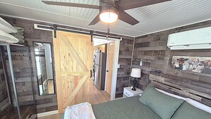 A bedroom with a bed, a nightstand, a lamp, a ceiling fan, and a wooden door. - Redbud Tiny Home Resort (Big Cabin)