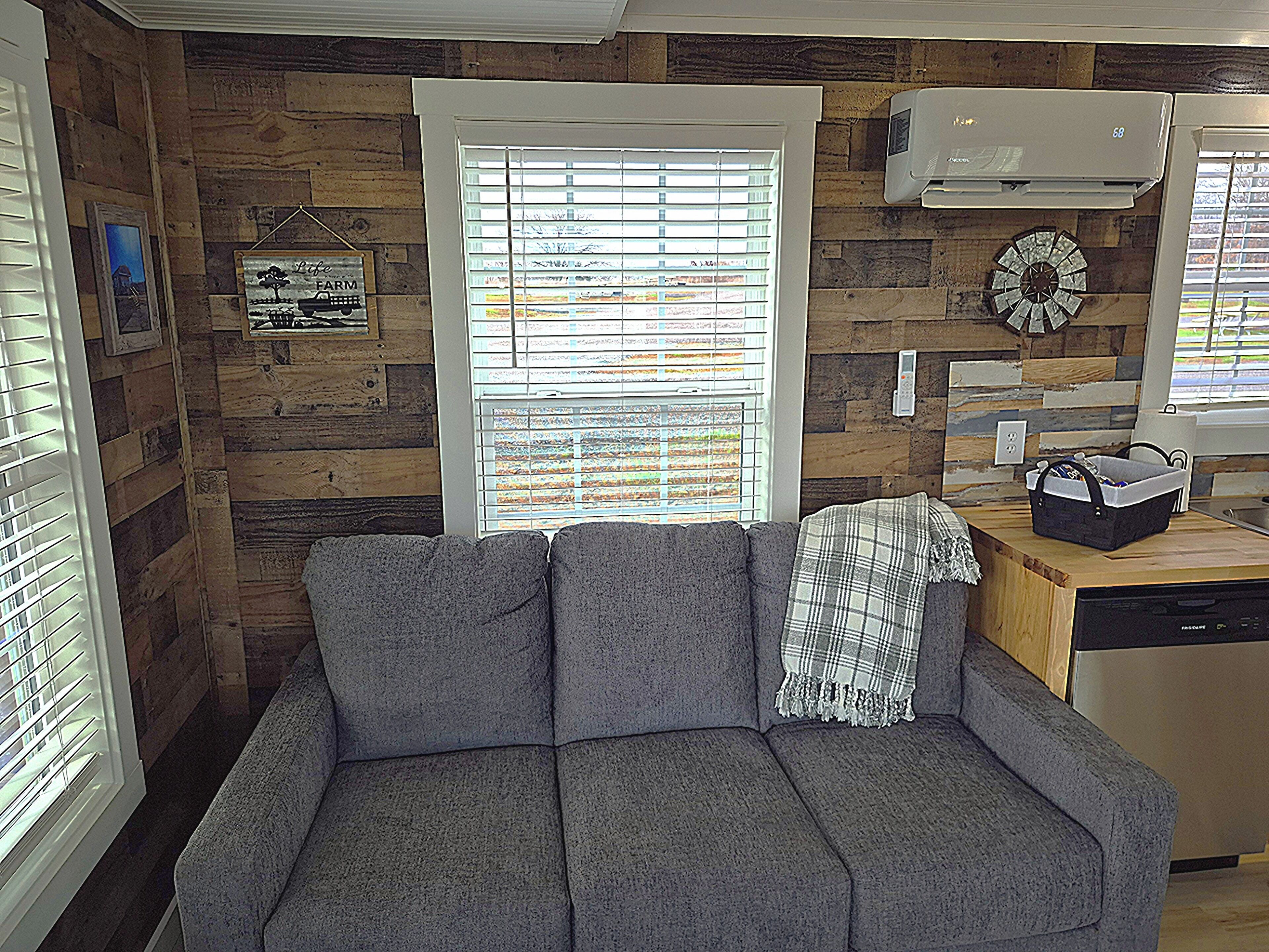 A cozy living area with a gray sofa, a wooden wall, a window with blinds, and a kitchen area with a dishwasher.