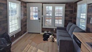 A room with a wooden floor, a gray sofa, a wooden dining table, and a white door with a window. - Redbud Tiny Home Resort (Big Cabin)